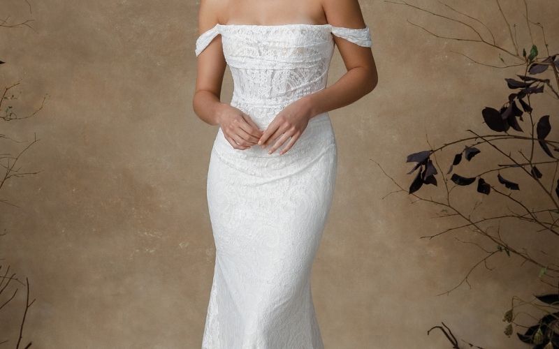 Bride wearing a trending off-the-shoulder lace wedding dress with a fitted silhouette and chapel train, standing against a soft neutral background